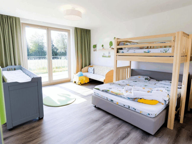 Helles Kinderzimmer mit großem Fenster, Etagenbett, grauem Kinderbett, kleiner Couch, grünen Vorhängen und buntem Dekor. Sonnenlicht füllt den Raum; verschiedene Bettarten sind zusammen mit einem grünen Teppich und Spielzeug-LKW auf dem Boden vorgestellt.