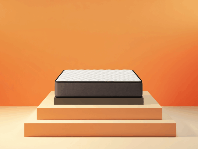 Eine Matratze mit weißer Steppdecke und dunklen Seiten wird auf einem dreistufigen Podest in Beige und Orange vor orangefarbenem Hintergrund präsentiert - ideal für Verbraucher, die Qualität suchen, die in Matratzen-Testergebnissen hervorgehoben wird.