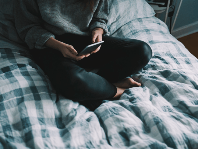 Eine Person sitzt im Schneidersitz auf einem Bett mit einer karierten Decke, trägt eine dunkle Hose und einen grauen Pullover, surft online auf ihrem Smartphone, um Matratzen zu kaufen, und blickt auf den Bildschirm.