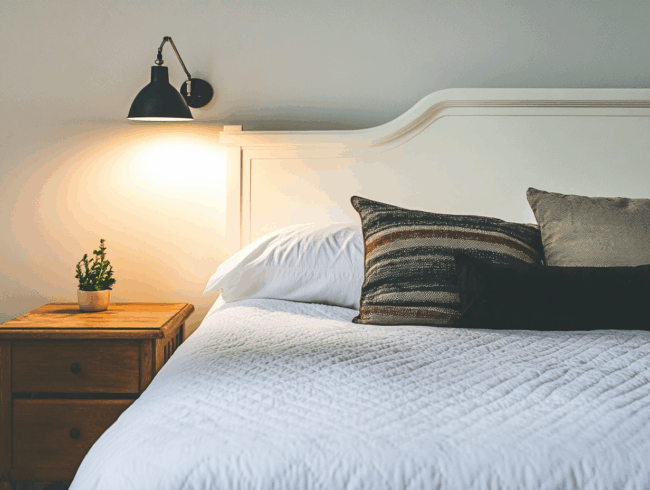 Ein ordentlich gemachtes Queensize-Bett mit weißer Bettwäsche, gestreiften und festen Kissen, steht neben einem hölzernen Nachttisch mit einer kleinen Topfpflanze, unter einer schwarzen Wandlampe, die warmes Licht spendet.