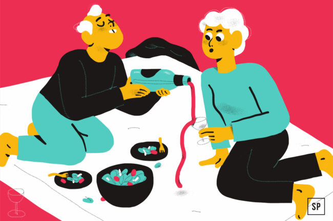 Zwei Personen mit weißen Haaren sitzen auf einem Matratzenschoner, wobei die eine dem anderen Rotwein in sein Glas gießt. In der Nähe stehen zwei Schalen mit Snacks vor einem leuchtend rosa-weißen Hintergrund.