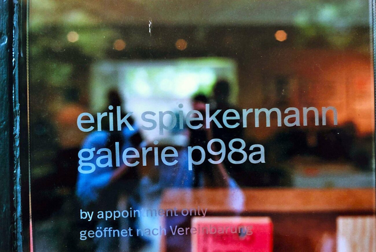 Nahaufnahme einer Glastür mit dem Text: erik spiekermann galerie p98a in Kleinbuchstaben und darunter nur nach Vereinbarung geöffnet, mit einer verschwommenen Innenraumszene, die sich im Glas spiegelt.