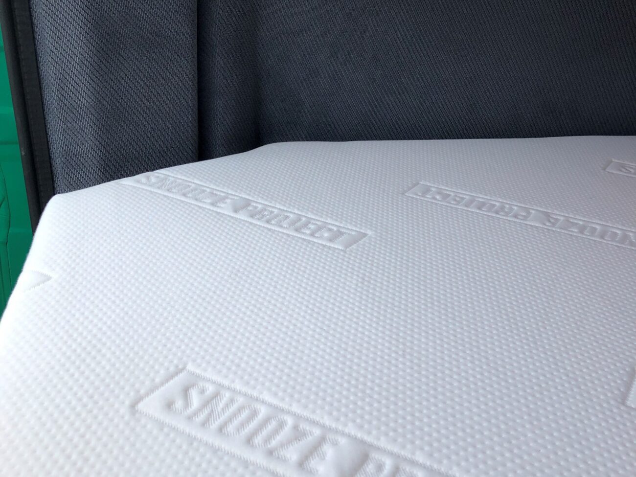 Nahaufnahme einer weißen Matratze mit strukturierter Oberfläche und geprägten SNOOZE PROJECT-Logos vor einem dunkelgrauen, gepolsterten Kopfteil.