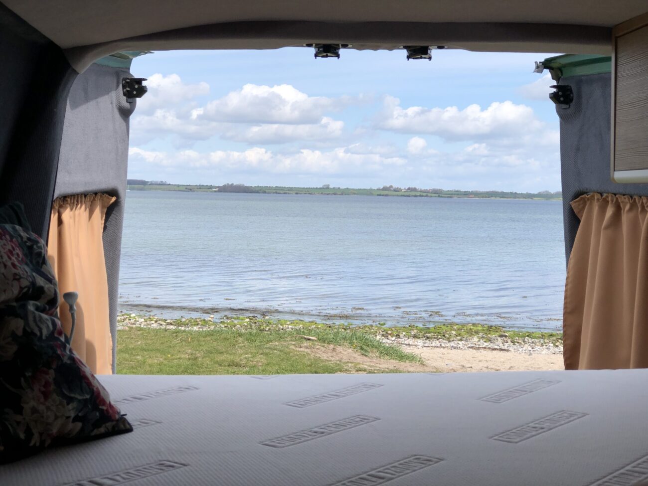 Blick aus dem Inneren eines Wohnmobils, der durch die offene Hintertür auf einen ruhigen See mit grasbewachsenem und felsigem Ufer unter einem teilweise bewölkten Himmel fällt. Im Vordergrund sind der Rand eines Bettes und Kissen zu sehen.