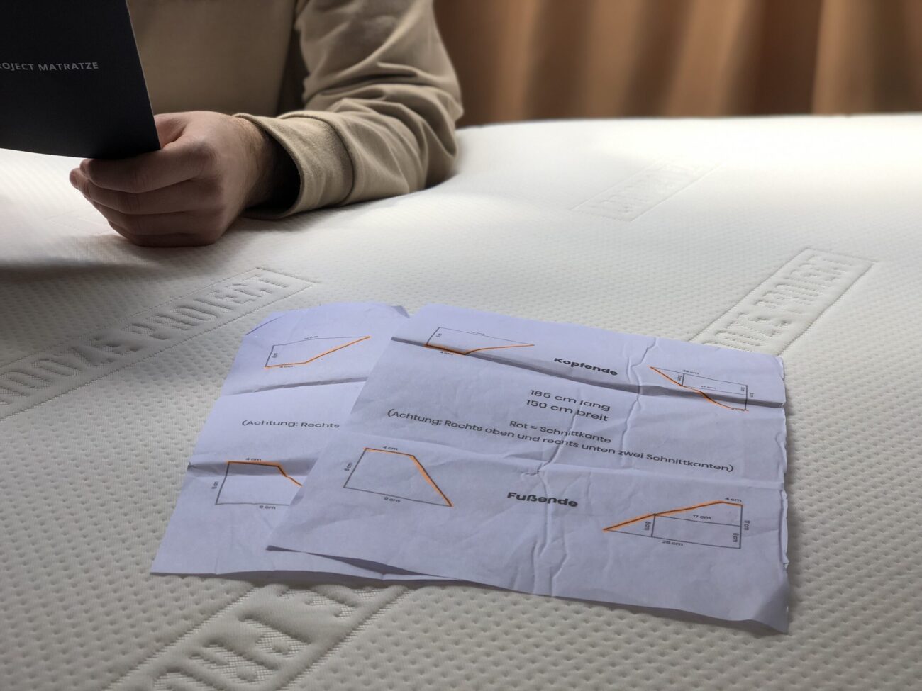 Eine Person hält ein schwarzes Heft über eine weiße Matratze. Auf der Matratze befinden sich zwei Blätter mit Diagrammen mit Abmessungen und Beschriftungen in deutscher Sprache bei natürlichem, weichem Licht.