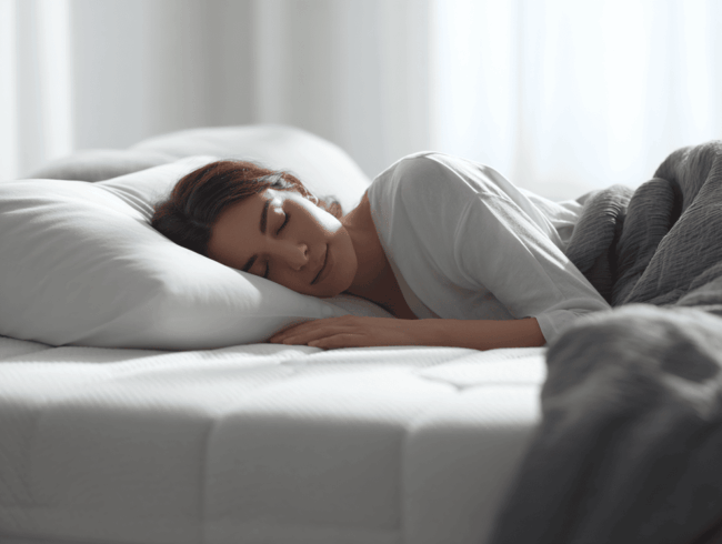 Eine Frau mit dunklem Haar schläft friedlich auf einem Bett, auf der Seite liegend, mit dem Kopf auf einem weißen Kissen, das für die Druckentlastung entworfen wurde und von einer grauen Decke bedeckt ist. Weiches natürliches Licht fällt durch die Vorhänge im Hintergrund.
