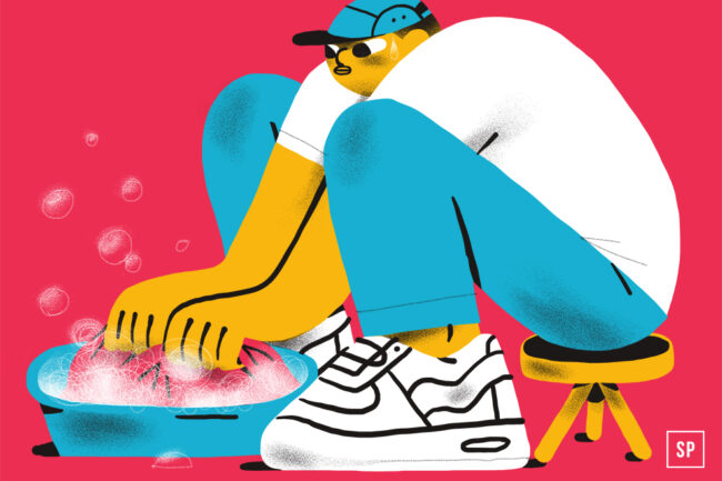 Eine Person mit Mütze, weißem Hemd, blauer Hose und Turnschuhen hockt auf einem Hocker und wäscht vor einem leuchtend rosa Hintergrund Kleidung in einem Seifeneimer.