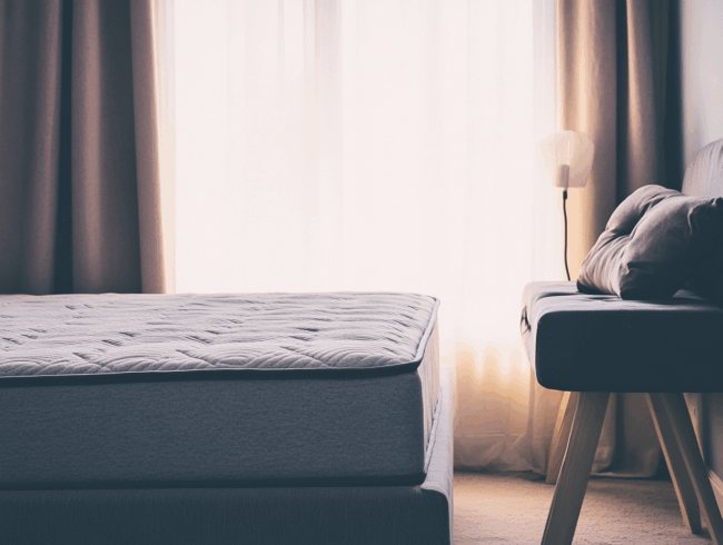 Ein ordentlich gemachtes Bett mit einer Matratze mit Stockflecken steht neben einem modernen grauen Stuhl mit Kissen am Fenster. Durchsichtige Vorhänge lassen sanftes natürliches Licht in den Raum, und eine hohe Lampe steht direkt hinter dem Stuhl.