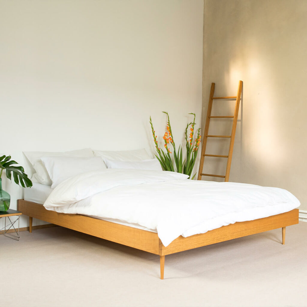 Minimalistisches Schlafzimmer mit einem hölzernen Bettgestell, weißer Bettwäsche, grünen Pflanzen und einer an die Wand gelehnten Holzleiter, alles auf einem hellen Teppich und neutralen Wänden.