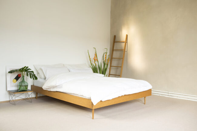 Ein minimalistisches Schlafzimmer mit einem Holzbettgestell, weißer Bettwäsche und weißen Wänden. Auf einem kleinen Beistelltisch steht eine grüne Vase mit Blättern, und hohe orangefarbene Blumen stehen neben einer Holzleiter, die an der Wand lehnt.