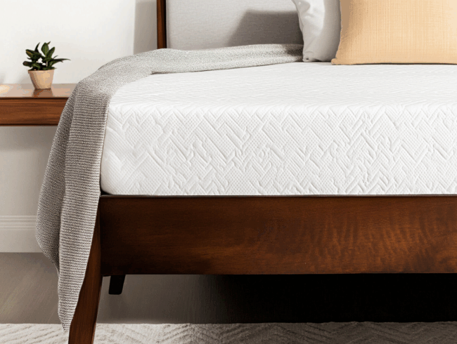 Nahaufnahme eines ordentlich gemachten Bettes mit einer strukturierten weißen Matratze, die dafür sorgt, dass die Matratze nicht rutscht, einem Holzrahmen, einer über die Seite drapierten grauen Decke und einer kleinen Topfpflanze auf dem hölzernen Nachttisch im Hintergrund.