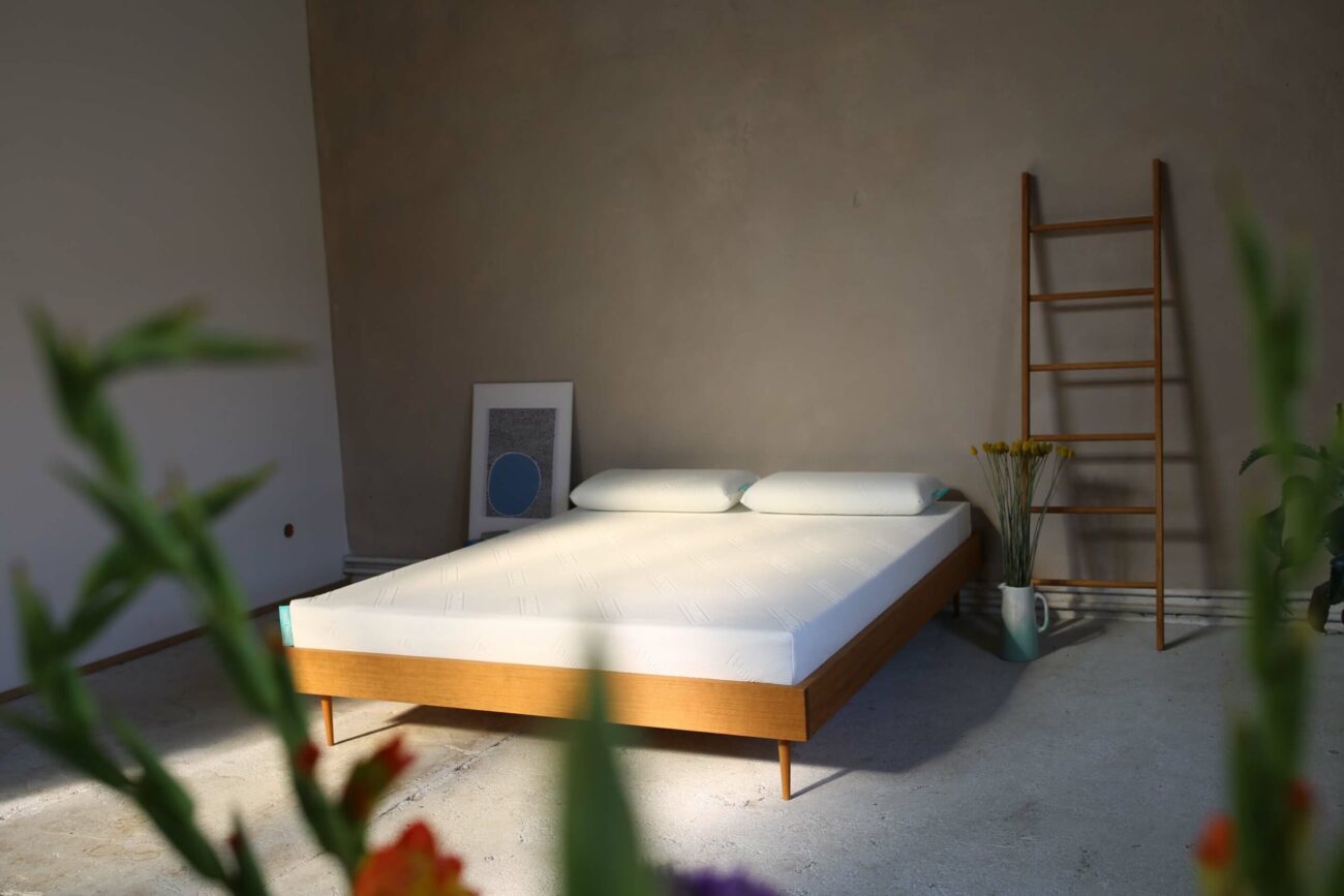 Ein minimalistisches Schlafzimmer mit einem Holzbettgestell, zwei Kissen und einer weißen Matratze. In der Ecke befinden sich eine Holzleiter, ein gerahmtes Kunstwerk, eine Vase mit gelben Blumen und unscharfe Pflanzen im Vordergrund.