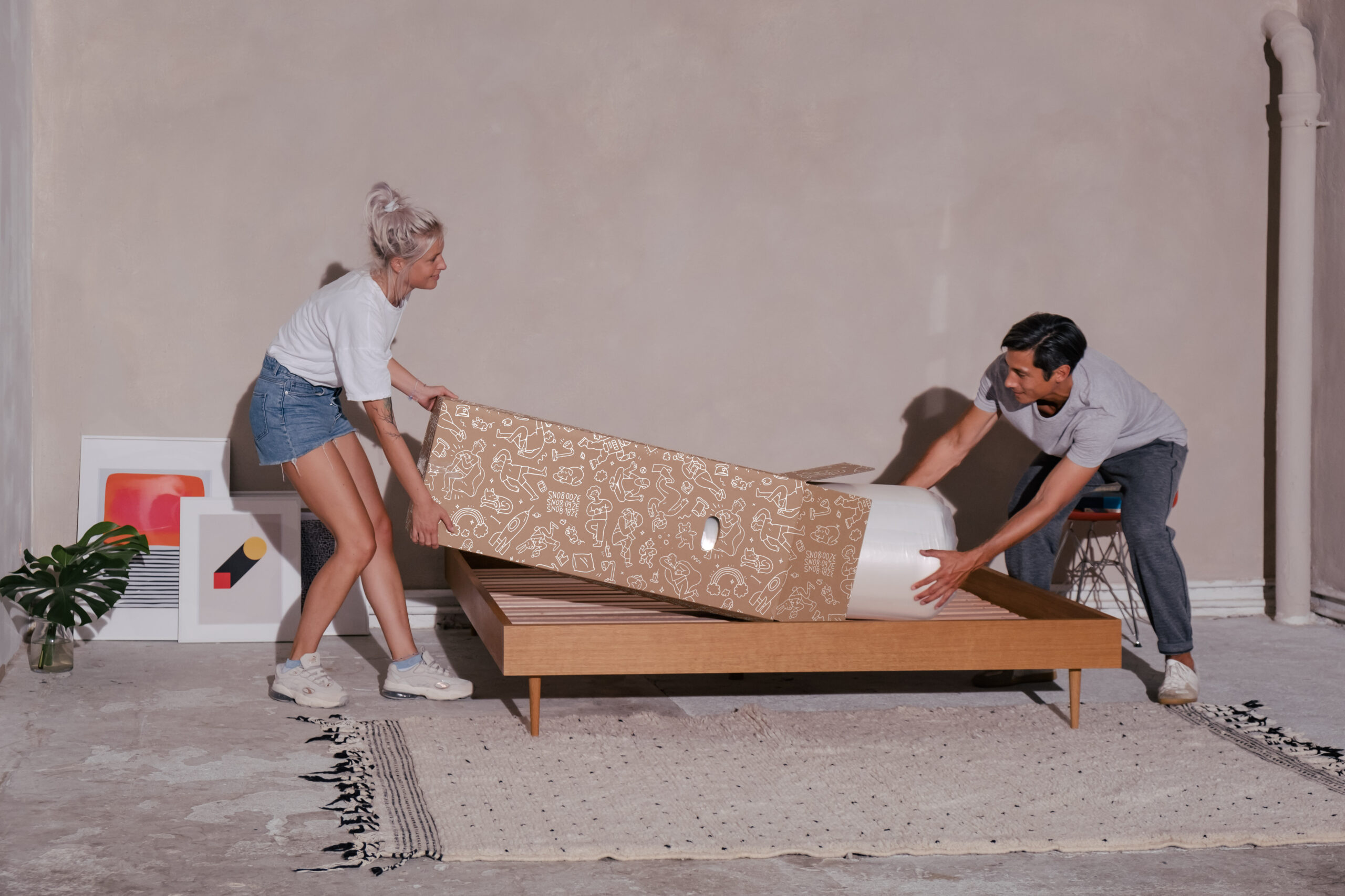 Eine Frau und ein Mann packen in einem minimalistischen Raum mit Kunstwerken auf dem Boden und einer Topfpflanze in der Nähe eine zusammengerollte Matratze auf ein hölzernes Bettgestell aus.