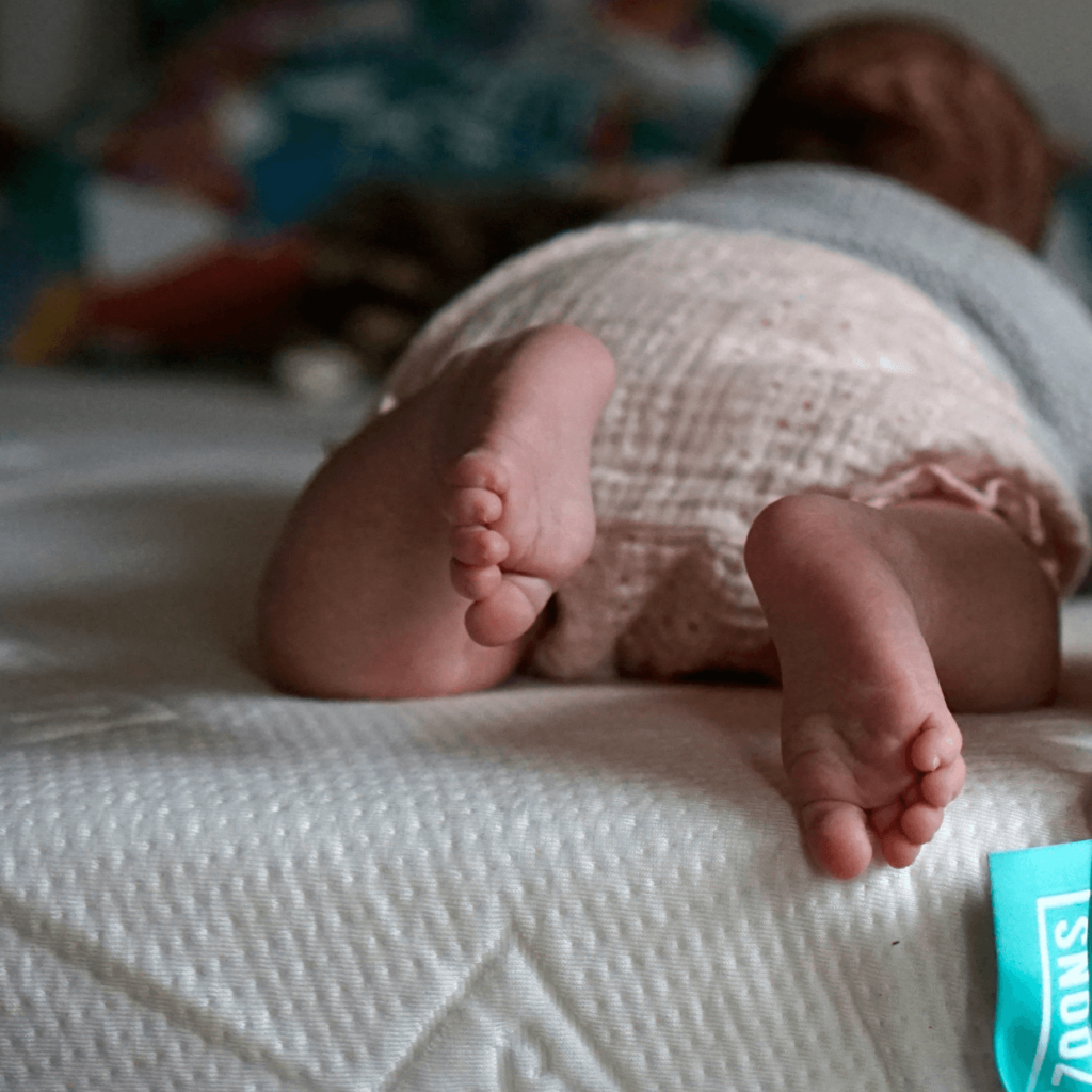 Ein Baby liegt auf dem Bauch auf einem Bett, trägt strukturierte Shorts und ein graues Oberteil, wobei seine nackten Füße und Beine im Mittelpunkt stehen. Der Hintergrund ist sanft verschwommen.