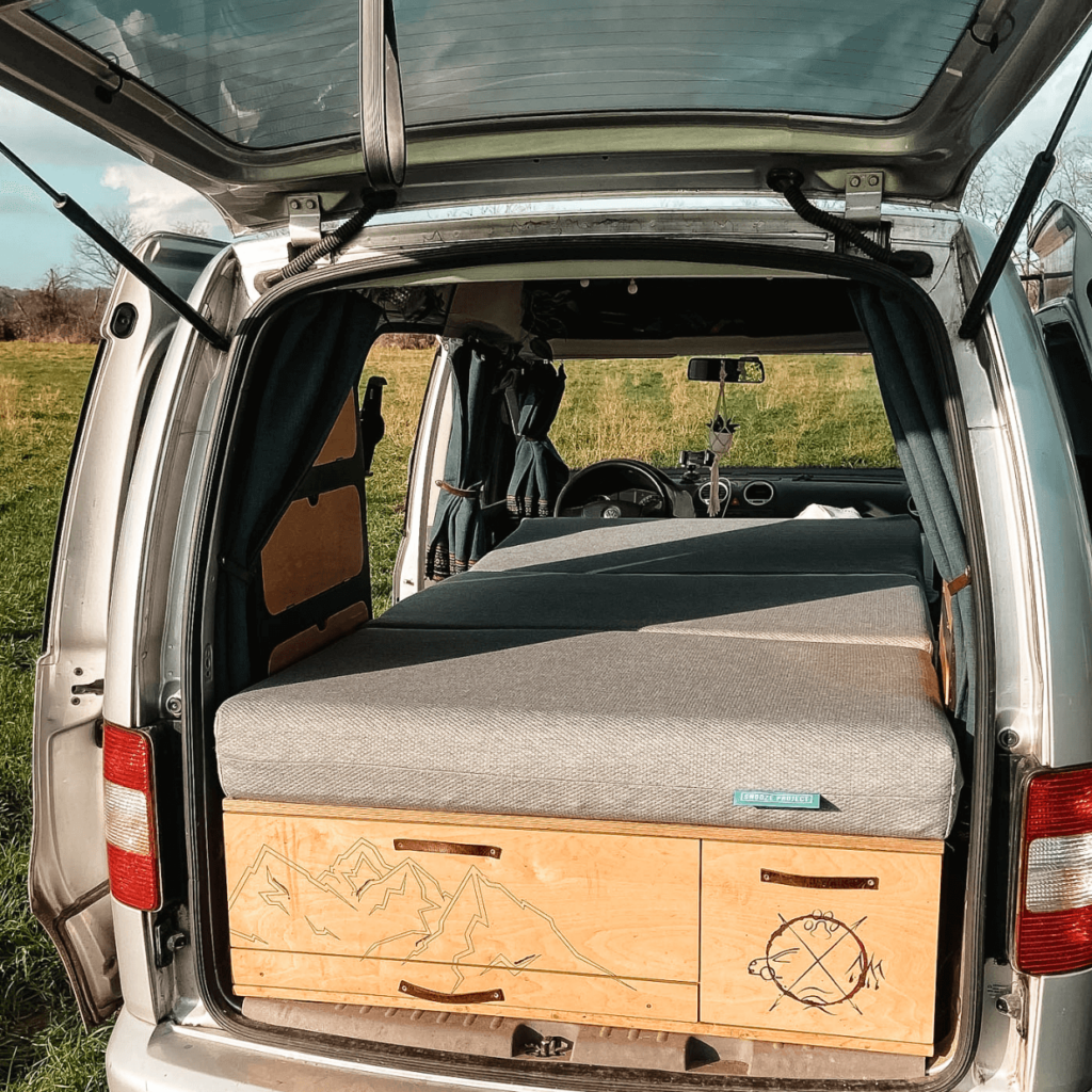 Ein Wohnmobil mit offener Hecktür, die ein ordentlich gemachtes Bett mit grauem Bettzeug und hölzernen Schubladen darunter zeigt, geparkt auf einer Wiese mit blauem Himmel im Hintergrund.