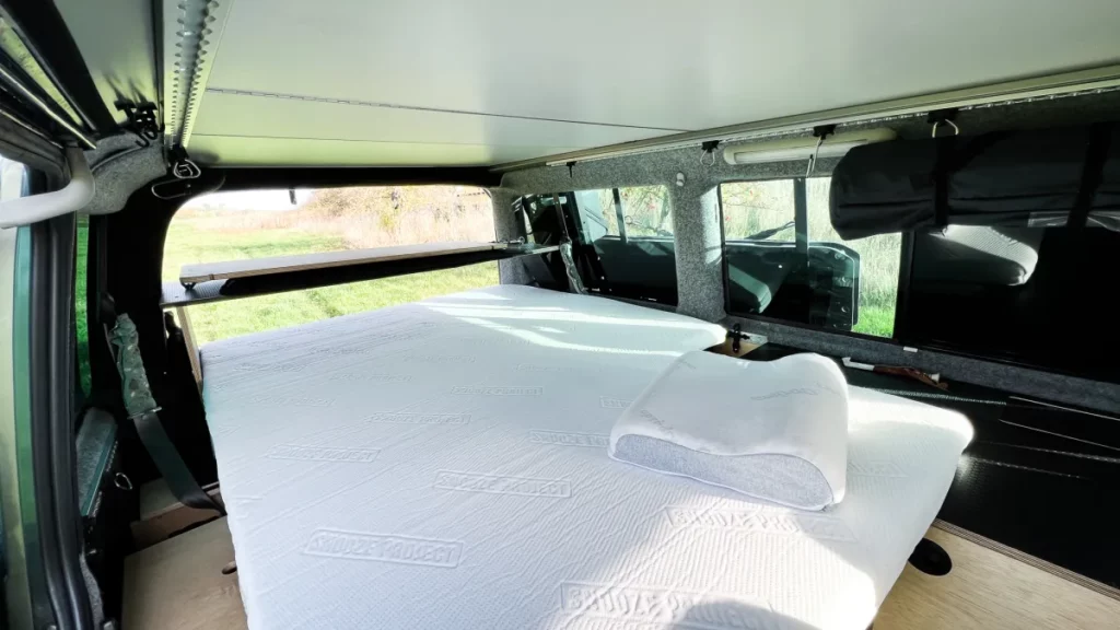 Ein ordentlich gemachtes Bett mit einem Kissen im Inneren eines Wohnmobils mit großen Fenstern, einfallendem Sonnenlicht und grünem Gras draußen. Der Innenraum scheint kompakt und effizient zum Schlafen eingerichtet zu sein.