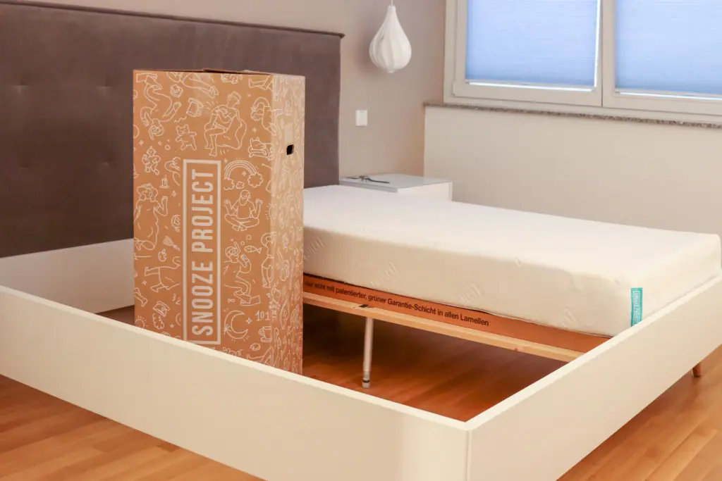 Eine verpackte Matratze mit der Aufschrift Snooze Project steht neben einer unverpackten Matratze auf einem weißen Bettgestell in einem modernen Schlafzimmer mit Holzboden und einem großen Fenster.