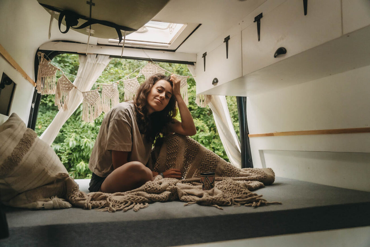 Eine Frau sitzt auf einem Bett in einem gemütlich eingerichteten Wohnmobil, umgeben von Decken und Kissen, während das Sonnenlicht durch eine Dachluke hereinfällt und Grünpflanzen vor der offenen Tür zu sehen sind.