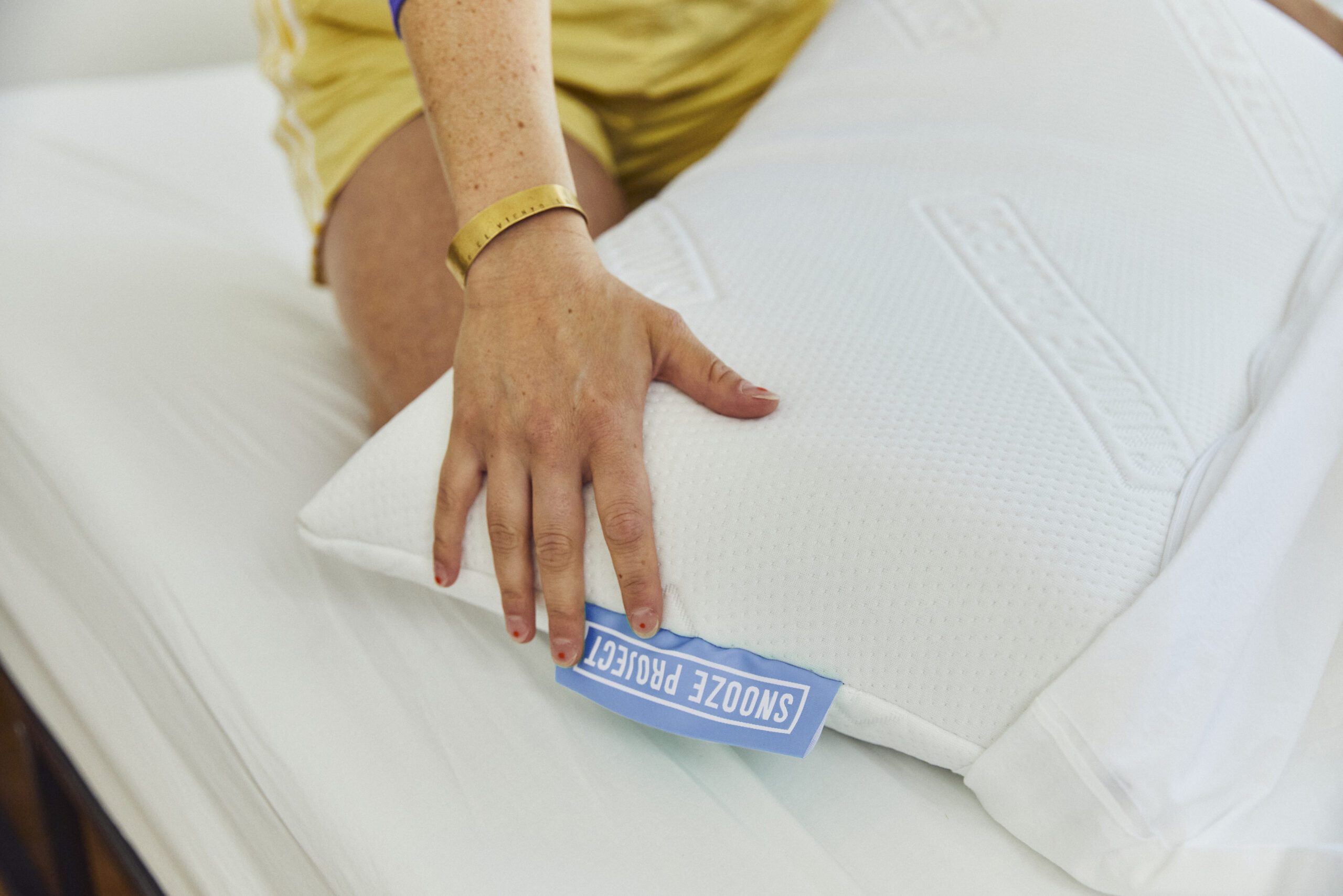Eine Person in einem gelben Outfit legt ein Kissen auf ein Bett. Auf dem Kissen, das mit einem blauen SNOOZE PROJECT-Tag gekennzeichnet ist, ist ihre linke Hand mit einem goldenen Armband und einem Ring zu sehen - ein Ausdruck des mühelosen Geistes von "Frei" im täglichen Leben.