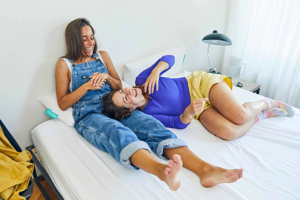 Zwei lächelnde und lachende Menschen auf einem Bett; einer sitzt mit gekreuzten Beinen, der andere liegt mit dem Kopf auf dem Schoß des Sitzenden. Der Raum ist hell und gemütlich und das Licht fällt durch ein Fenster.