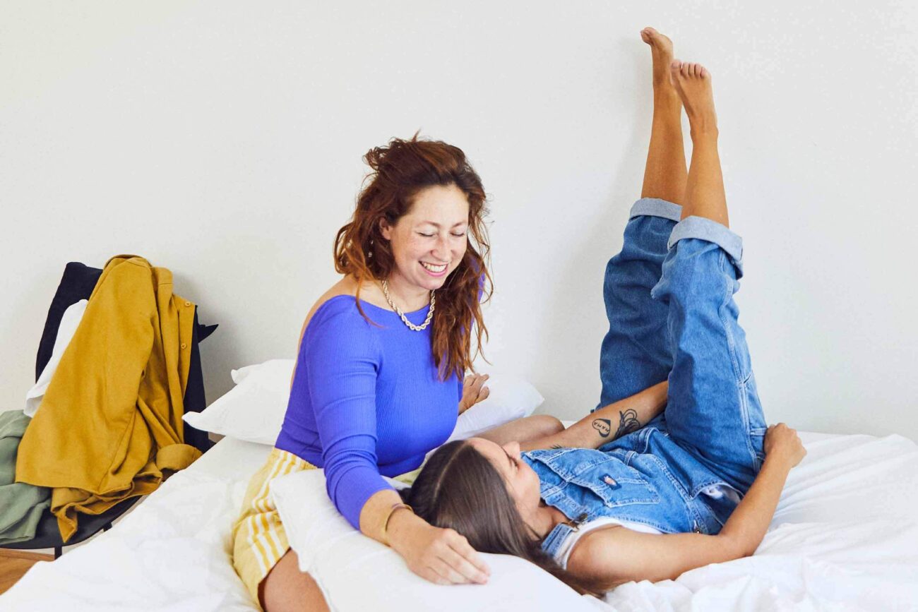 Zwei Frauen entspannen sich auf einem Bett, lächeln und lachen zusammen. Die eine sitzt in einem blauen Top, die andere liegt mit den Beinen an der Wand und trägt einen Jeans-Overall. Ein gelber Mantel hängt an einem Stuhl im Hintergrund.