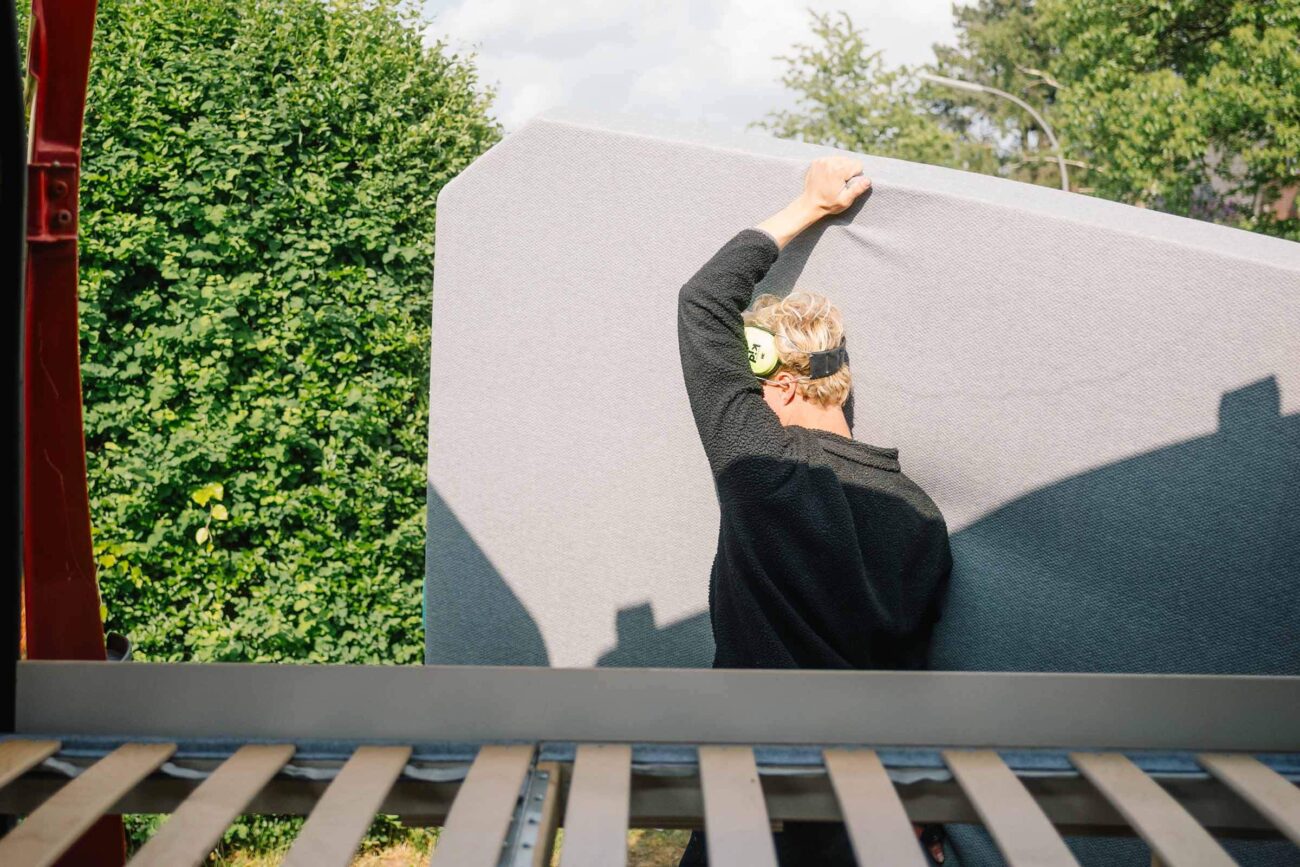 Eine Person mit Schutzbrille hebt eine große graue Matratze oder Platte von einem Fahrzeug, im Hintergrund sind grüne Büsche und Bäume zu sehen.