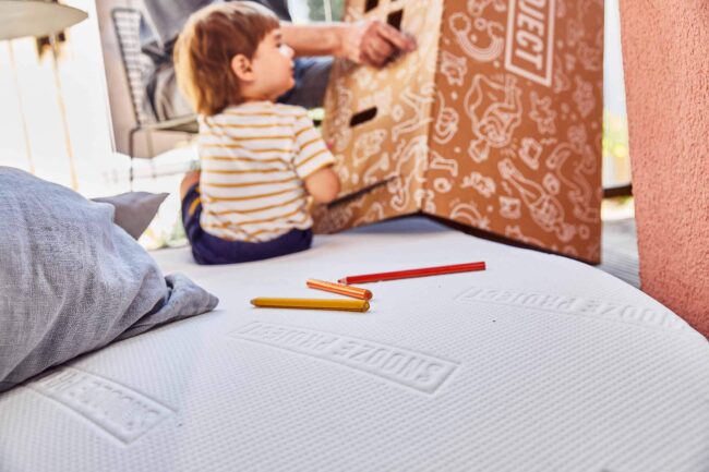 Ein kleines Kind in einem gestreiften Hemd sitzt auf dem Boden in der Nähe einer großen, verzierten Pappschachtel, mit Buntstiften und einem Kissen auf einer weißen, strukturierten Fläche im Vordergrund.