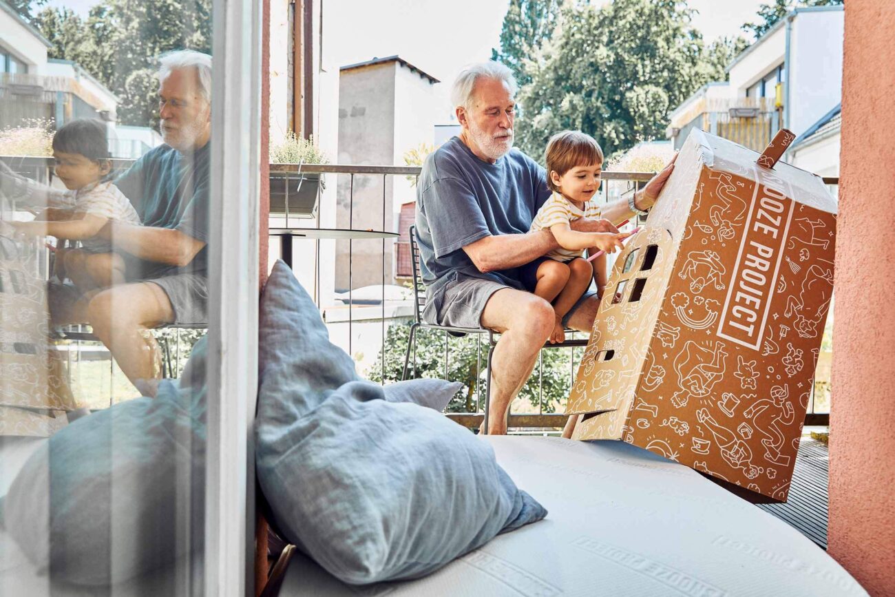 Ein älterer Mann sitzt auf einem Balkon mit einem kleinen Kind auf seinem Schoß. Sie interagieren mit einem großen, illustrierten Pappkarton. Ihr Spiegelbild erscheint in der Glastür nebenan. Im Vordergrund befinden sich Kissen.