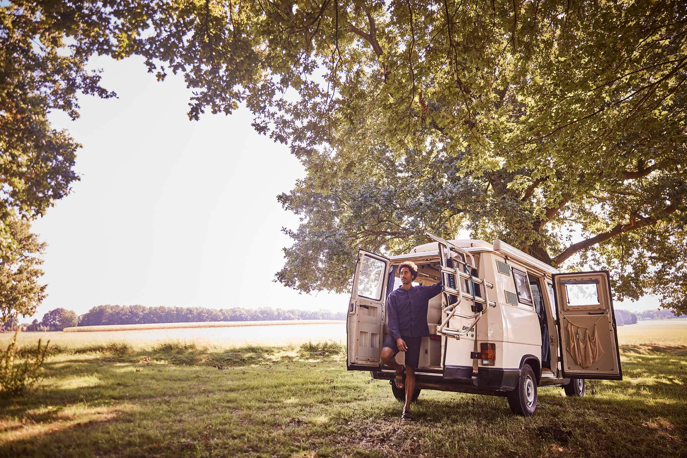 Eine Person steht in der offenen Tür eines beigen Wohnmobils, das unter einem großen Baum auf einer sonnigen, grasbewachsenen Wiese geparkt ist, und genießt die freie Natur.