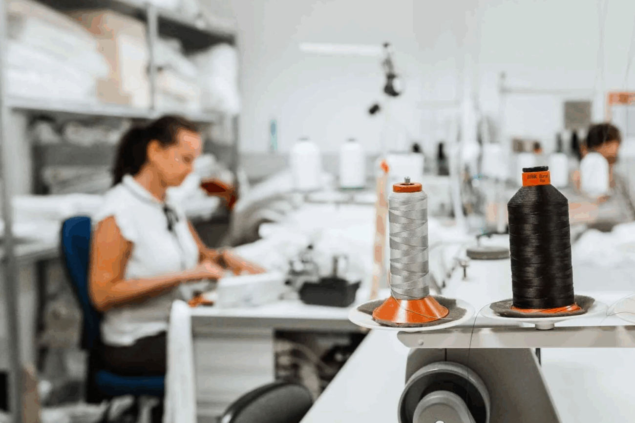 Zwei große Garnspulen, eine graue und eine schwarze, stehen auf einem Tisch in einer Nähwerkstatt im Mittelpunkt. Im unscharfen Hintergrund arbeitet eine Frau an einer Nähmaschine - ein Einblick in Wer wir sind: kreativ und frei in unserem Handwerk.
