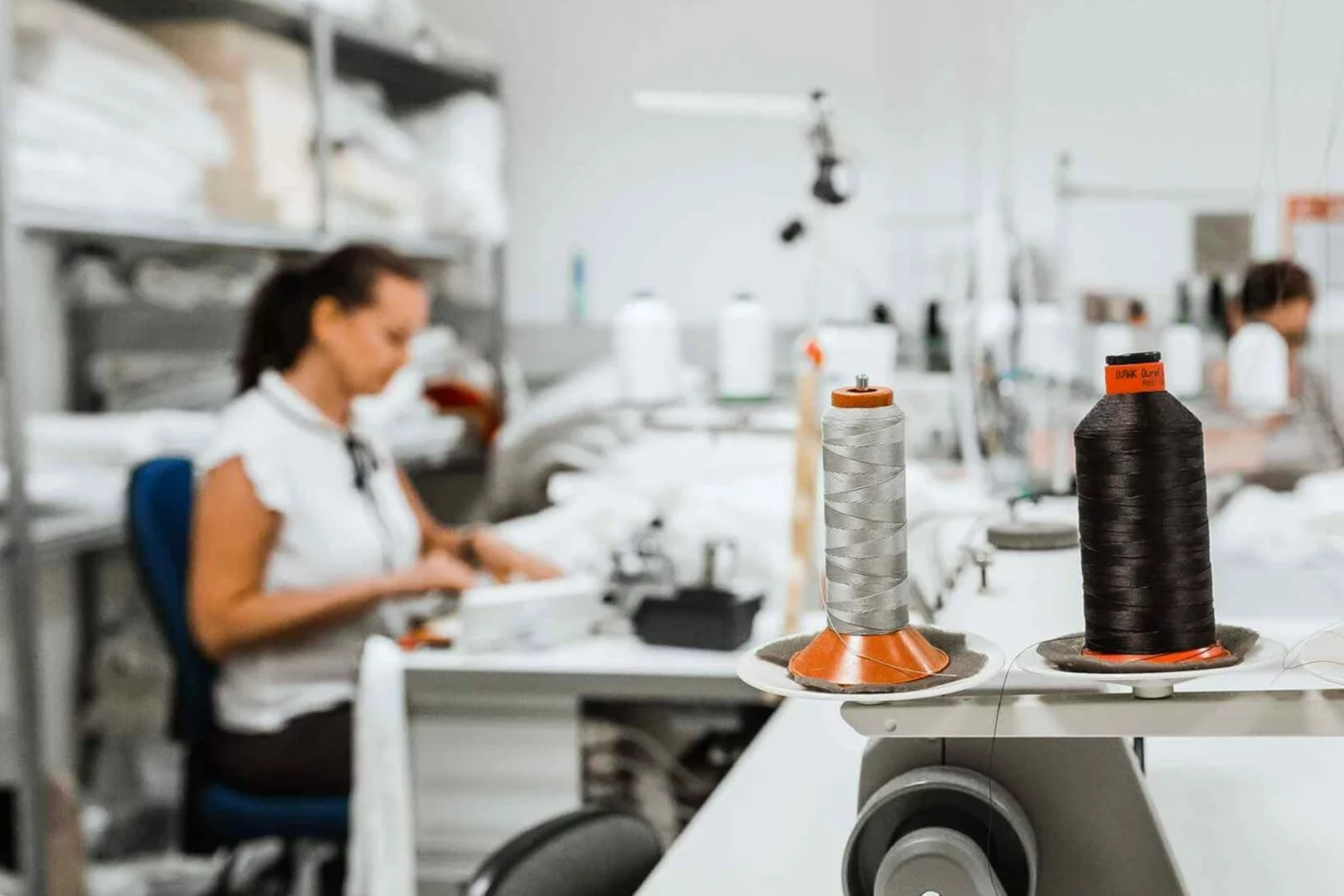 Zwei große Garnspulen, eine graue und eine schwarze, stehen auf einem Tisch in einer Nähwerkstatt im Mittelpunkt. Im unscharfen Hintergrund arbeitet eine Frau an einer Nähmaschine - ein Einblick in Wer wir sind: kreativ und frei in unserem Handwerk.