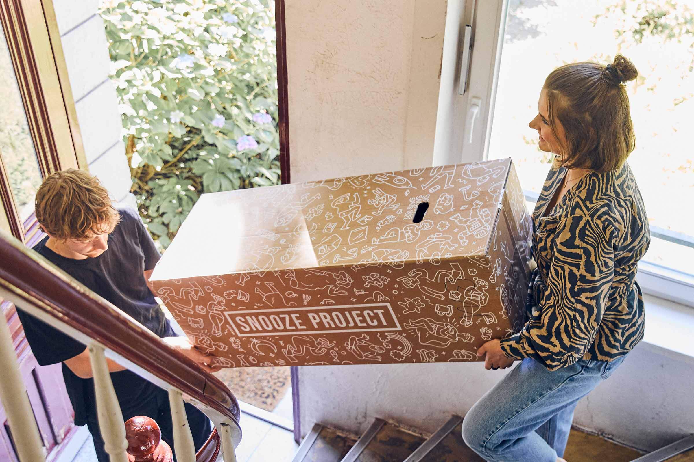 Zwei Personen tragen eine große Pappschachtel mit der Aufschrift "Snooze Project" eine Treppe hinauf, während das Sonnenlicht durch ein nahe gelegenes Fenster fällt und draußen das Grün der Bäume zu sehen ist.