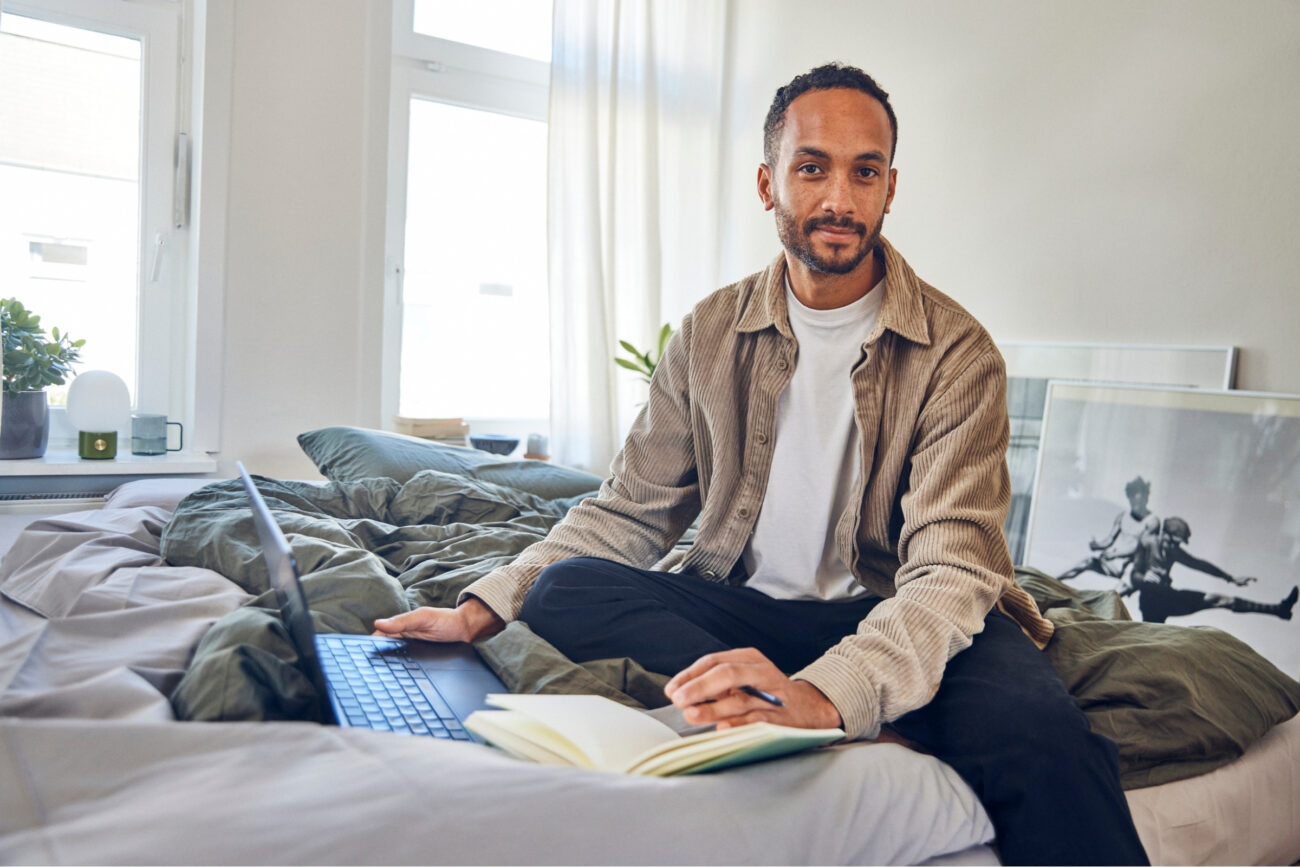 Ein Mann sitzt im Schneidersitz auf einem Bett mit einem Laptop und einem aufgeschlagenen Notizbuch, schreibt und schaut in die Kamera - vielleicht arbeitet er an Kundenbewertungen. Der helle Raum hat große Fenster, einige Pflanzen und ein gerahmtes Bild, das an der Wand lehnt.