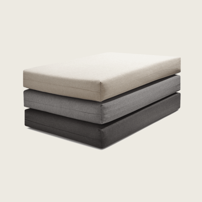 Ein Stapel von drei rechteckigen Kissen in Beige, Mittelgrau und Dunkelgrau, die vor einem schlichten hellen Hintergrund ordentlich übereinander angeordnet sind.