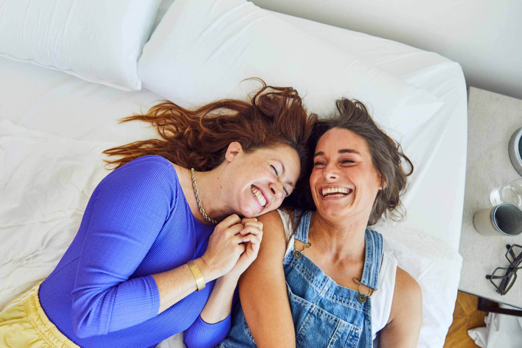 Zwei Frauen liegen auf einem Bett, lächeln und lachen zusammen. Die eine trägt ein blaues Oberteil, die andere einen Jeans-Overall. Sie halten sich an den Händen und wirken glücklich und entspannt.