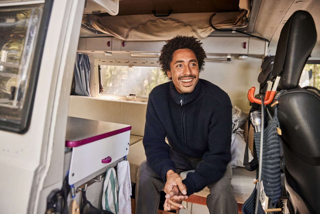 Ein Mann mit lockigem Haar und einem dunklen Pullover sitzt lächelnd in einem Wohnmobil. Dickere Polster und zugeschnittene Elemente verleihen dem organisierten Innenraum mit einer kleinen Theke, Handtüchern und hängenden Gegenständen Komfort und Stil.