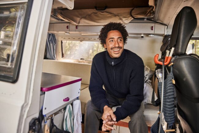 Ein Mann mit lockigem Haar und einem dunklen Pullover sitzt lächelnd in einem Wohnmobil. Dickere Polster und zugeschnittene Elemente verleihen dem organisierten Innenraum mit einer kleinen Theke, Handtüchern und hängenden Gegenständen Komfort und Stil.