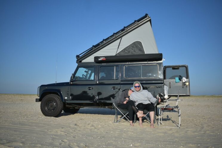 Nils und Kira sitzen in Campingstühlen vor ihrem Defender. 