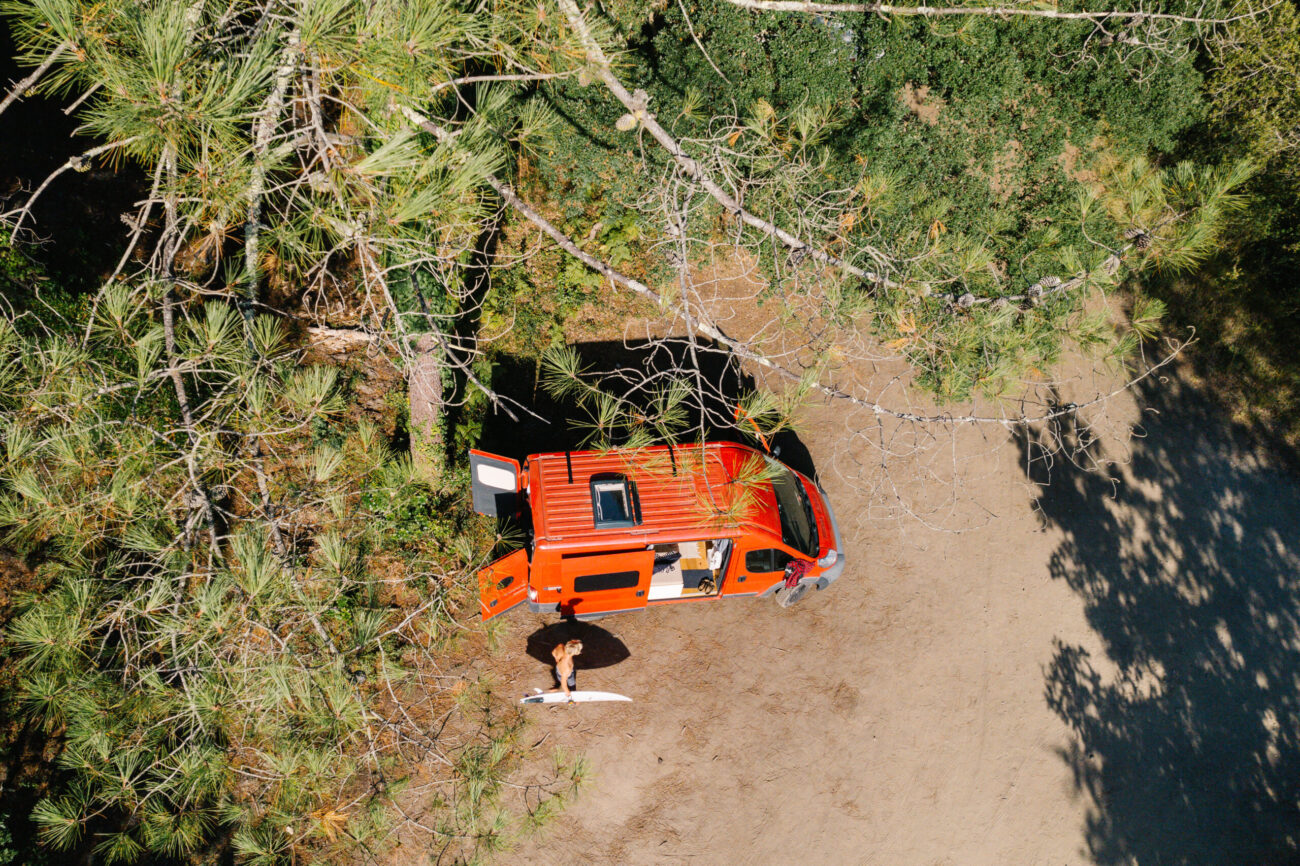 Luftaufnahme eines roten Busumbaus, der auf einer von Bäumen umgebenen, unbefestigten Lichtung geparkt ist. Eine Person steht außerhalb des Busses und hält ein Surfbrett, bereit für Abenteuer, während das Sonnenlicht Schatten auf den Boden wirft.