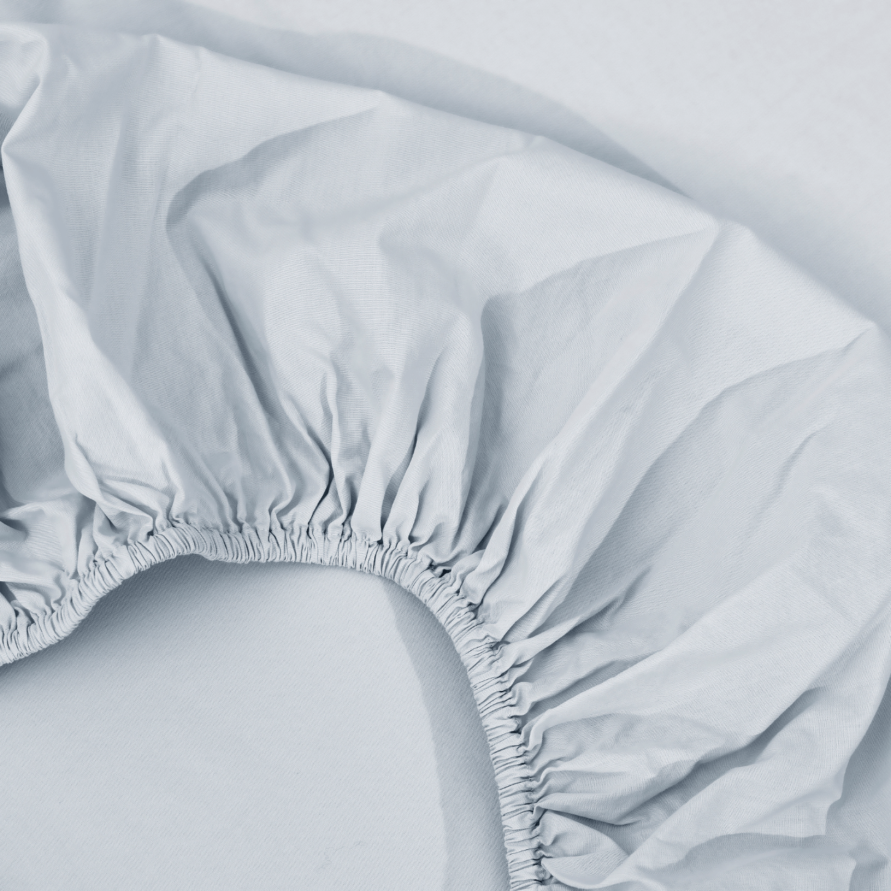 spannbettlaken nach maß hellgrau 2 Nahaufnahme eines hellblauen Spannbettlakens mit gerafften Gummirändern, das auf einer passenden blauen Stoffunterlage liegt.