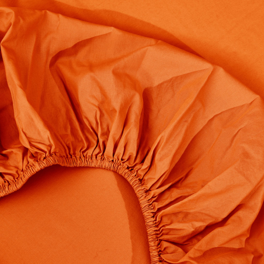 Eine Nahaufnahme eines orangefarbenen Spannbettlakens mit sichtbaren elastischen Rändern, das auf einer orangefarbenen flachen Oberfläche ausgebreitet ist.