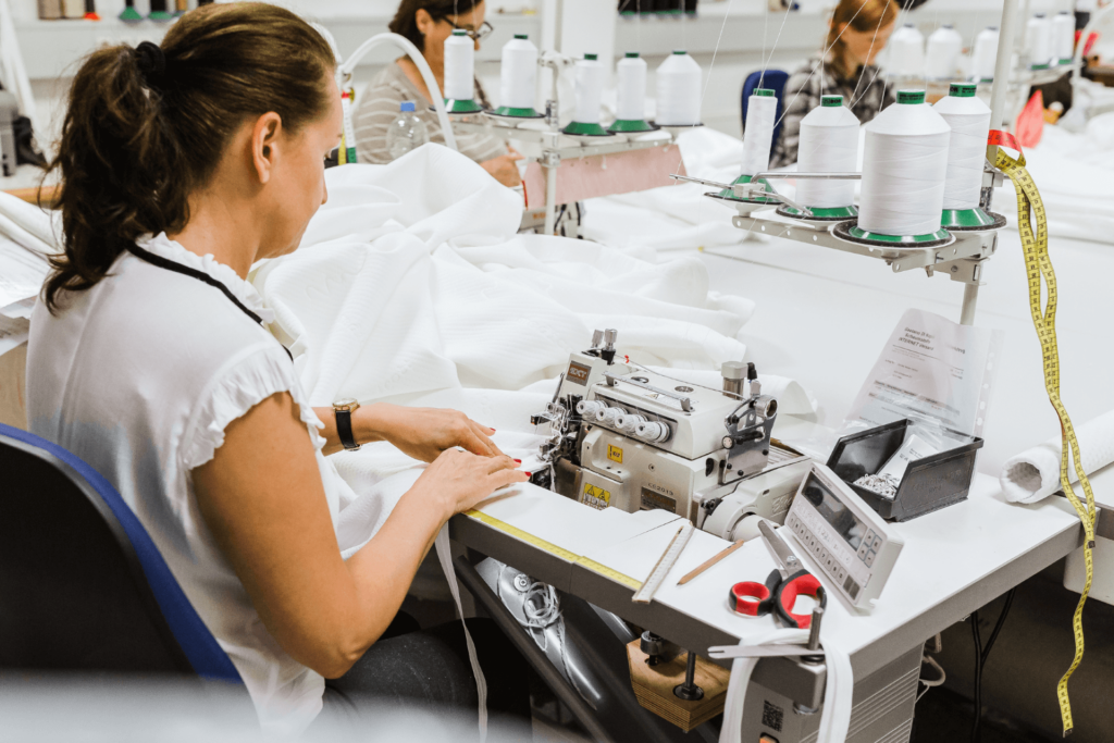 Eine Frau bedient eine Nähmaschine an einem Arbeitsplatz mit weißen Garnspulen, Stoff, Schere und Maßband in einer hellen, gut organisierten Bekleidungswerkstatt. Im Hintergrund sind andere Arbeiter beim Nähen zu sehen.