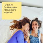 Zwei Frauen liegen auf einem Bett, lächeln und sehen glücklich aus. Der deutsche Text in einem gelben Kasten lautet: Für dich im Familienbetrieb in Deutschland hergestellt (Made for you in Germany).