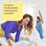 Zwei Frauen lächeln und machen zusammen ein Bett in einem hellen Raum. Auf einem gelben Kasten mit deutschem Text steht "Für dich im Familienbetrieb in Deutschland hergestellt".