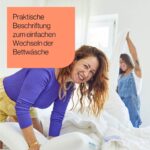 Zwei Frauen lächeln und wechseln die Bettwäsche in einem hellen Zimmer. Eine Frau trägt ein lila Oberteil, die andere einen Jeans-Overall. Der Text auf einer orangefarbenen Folie lautet: "Praktische Beschriftung zum einfachen Wechseln der Bettwäsche.".
