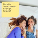 Zwei Frauen liegen auf einem Bett, lächeln und schauen sich an. Die eine trägt ein blaues Oberteil, die andere einen Jeans-Overall. In einem gelben Kasten in der Ecke steht ein deutscher Text: Für dich im Traditionsbetrieb in Portugal hergestellt.