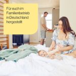 Eine lächelnde Frau spielt mit einem Baby auf einem Bett in einem hellen Raum, während ein Mann im Hintergrund in der Küche steht. Der deutsche Text auf dem gelben Kasten lautet: Für euch im Familienbetrieb in Deutschland hergestellt.