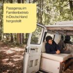 Ein Mann entspannt sich und lächelt, während er in einem Wohnmobil liegt, das in einem sonnenbeschienenen Wald geparkt ist. Der deutsche Text auf einem gelben Kasten lautet: Passgenau im Familienbetrieb in Deutschland hergestellt.