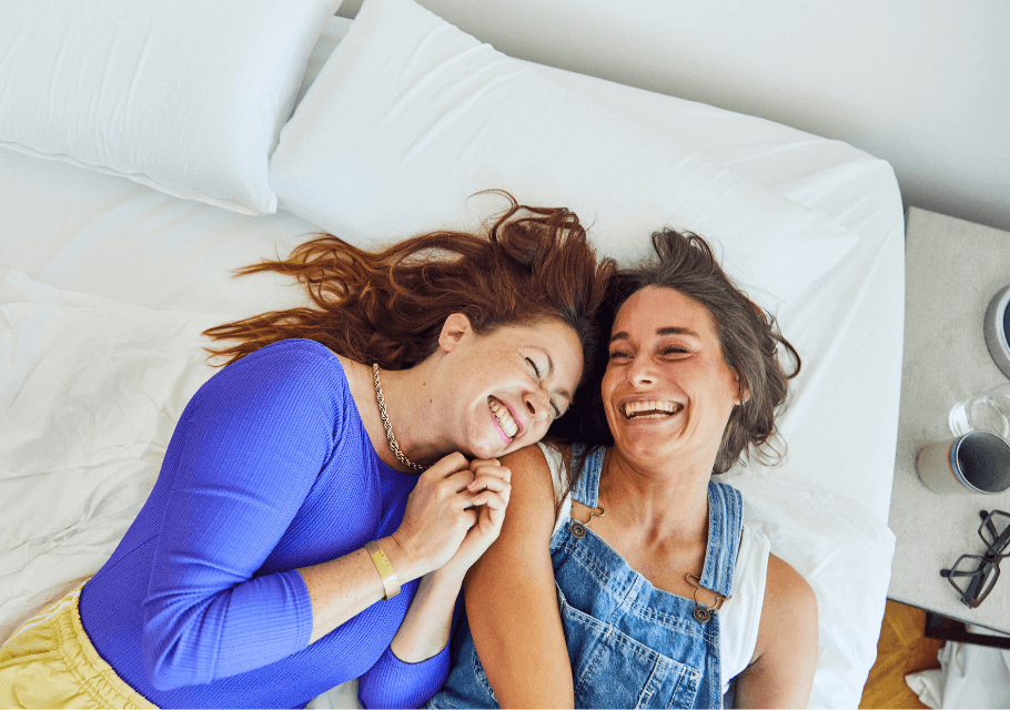 Zwei Frauen liegen Seite an Seite auf einem Bett, lächeln und lachen zusammen, umgeben von weißen Kissen und Decken. Eine Frau trägt ein blaues Oberteil, die andere einen Jeans-Overall.