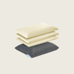 Ein Kissen aus Memory-Schaumstoff, dessen Schichten getrennt und darüber gestapelt sind, mit drei cremefarbenen Schaumstoffschichten und einem dunkelgrauen Stoffbezug mit einem kleinen blauen Etikett auf einem schlichten hellen Hintergrund.